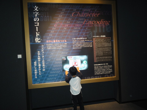 A little child playing an encodings game in front of a large poster about encodings.